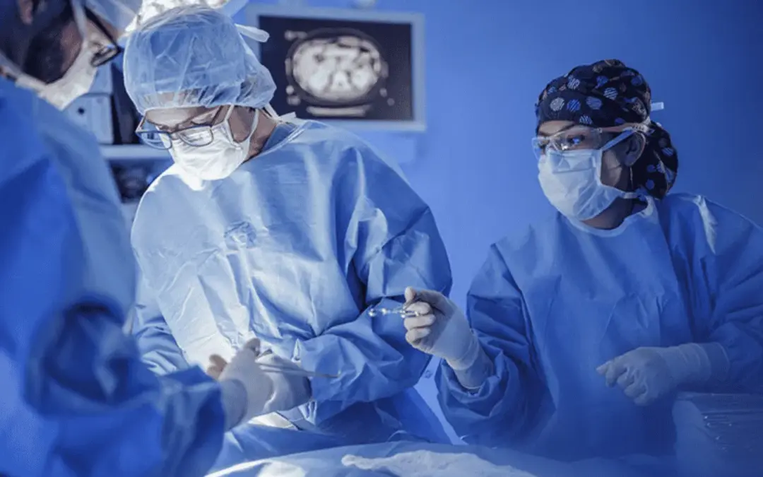 Surgeons in sterile gowns and masks performing surgery under bright operating room lights with a monitor in the background.