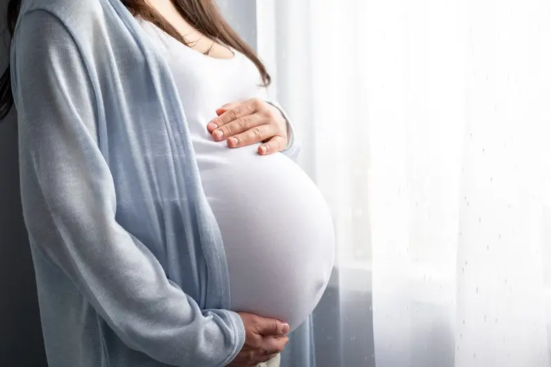 Pregnant woman standing by a window, wearing a light blue cardigan, hands resting on her rounded baby bump.