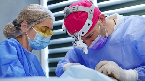 Picture Two surgeons in blue scrubs, masks, and headlamps perform surgery under bright operating lights, focused on the patient.