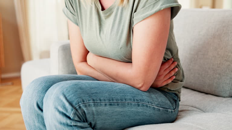 Person sitting on a gray sofa with arms wrapped around their abdomen, wearing a green T-shirt and blue jeans, suggesting stomach discomfort.