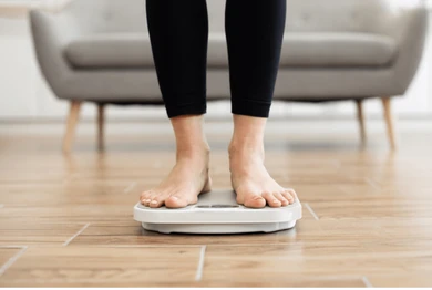 Person standing on a digital bathroom scale on a wooden floor in a living room silhouette style.
