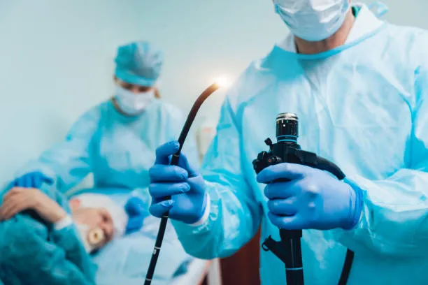 Oral cancer Surgeons in blue scrubs and masks performing surgery; one holds a handheld endoscope near a glowing cautery tool while a patient lies on the operating table in the background.