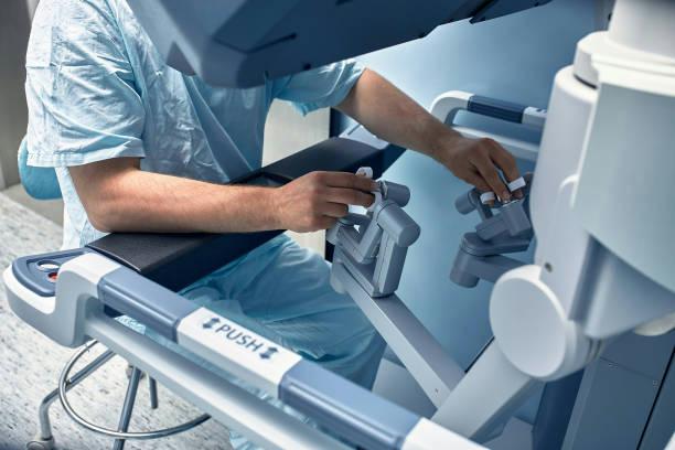 robotic thyroid surgery Medical professional in blue scrubs operating a robotic surgical console, manipulating levers and controls beside a patient bed and screen.